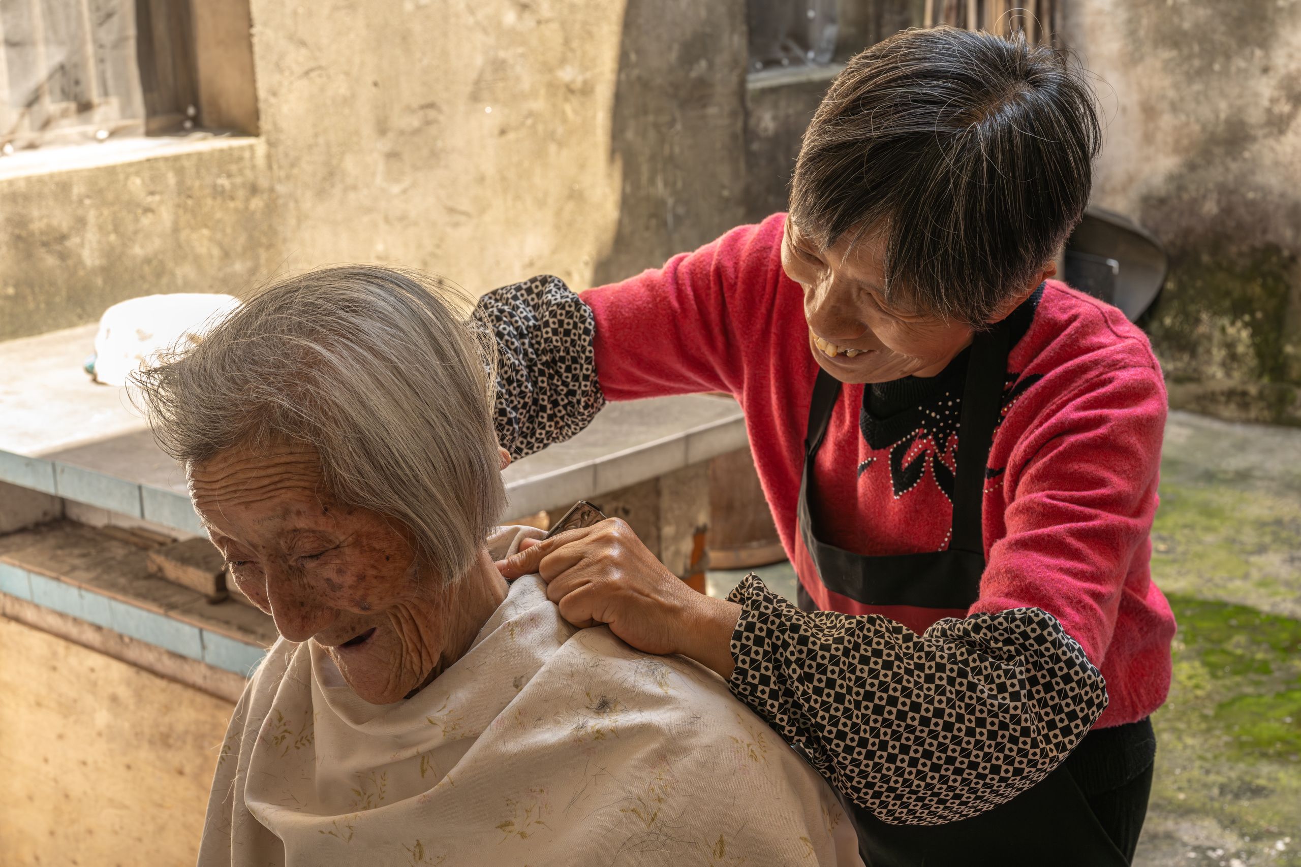 An elderly woman receives a haircut from another woman in a rustic outdoor setting.