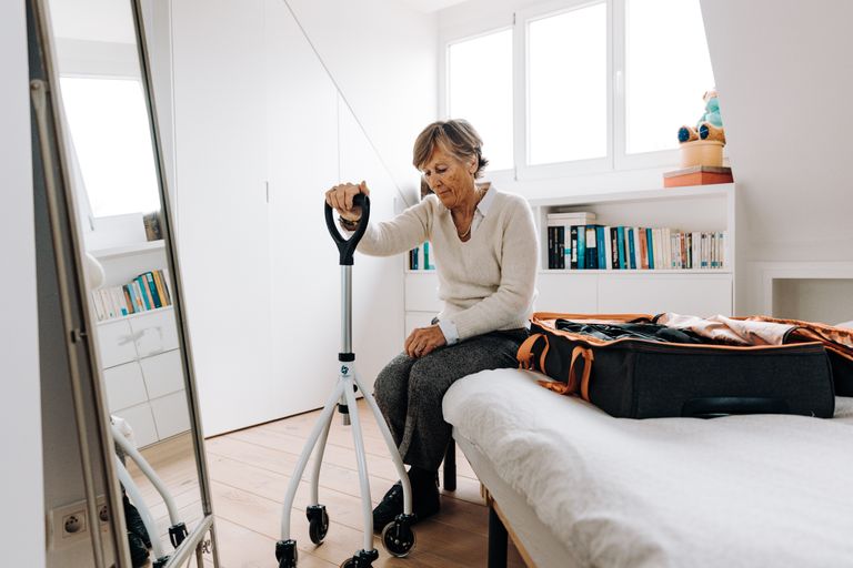 Senior woman sitting on bed with walker, preparing suitcase in bright bedroom.