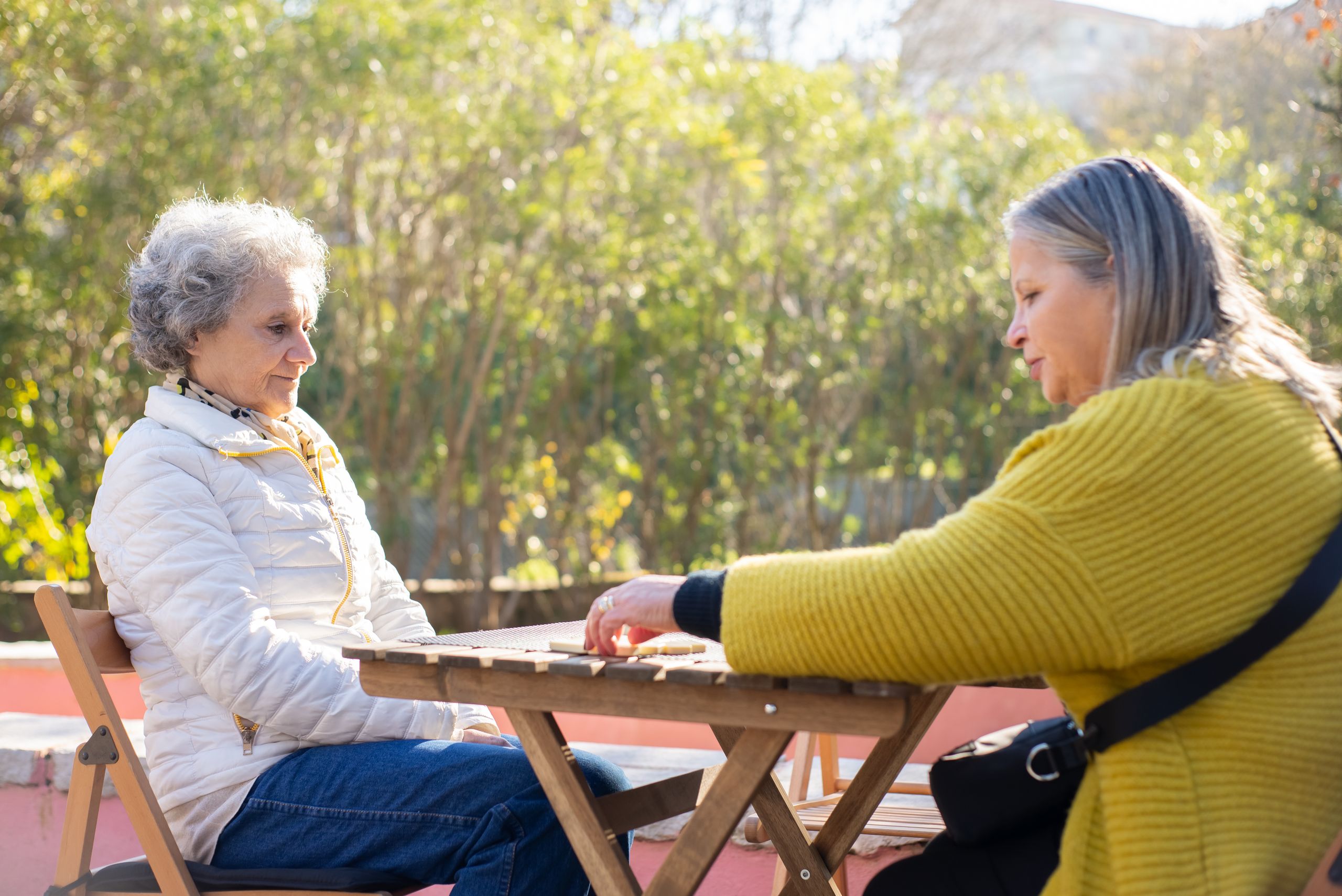 Two senior women enjoying a game of dominoes at a wooden table outdoors in sunny Portugal.