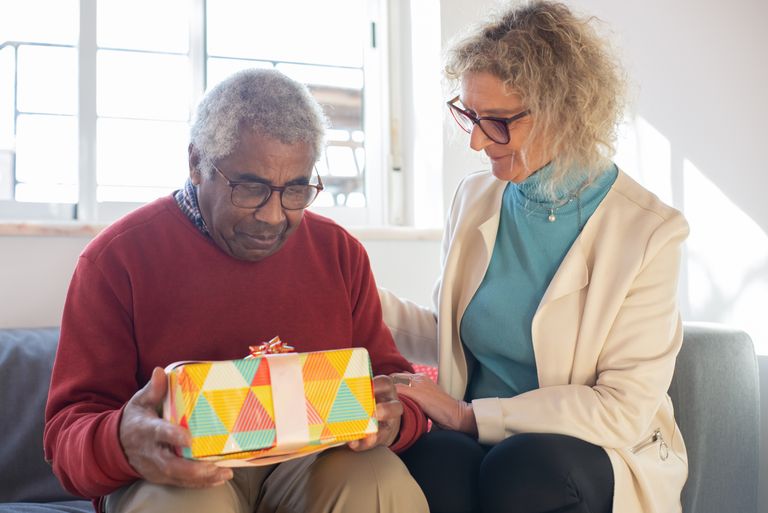 A senior man receives a colorful gift from a caring woman on a couch, indoors with natural light.