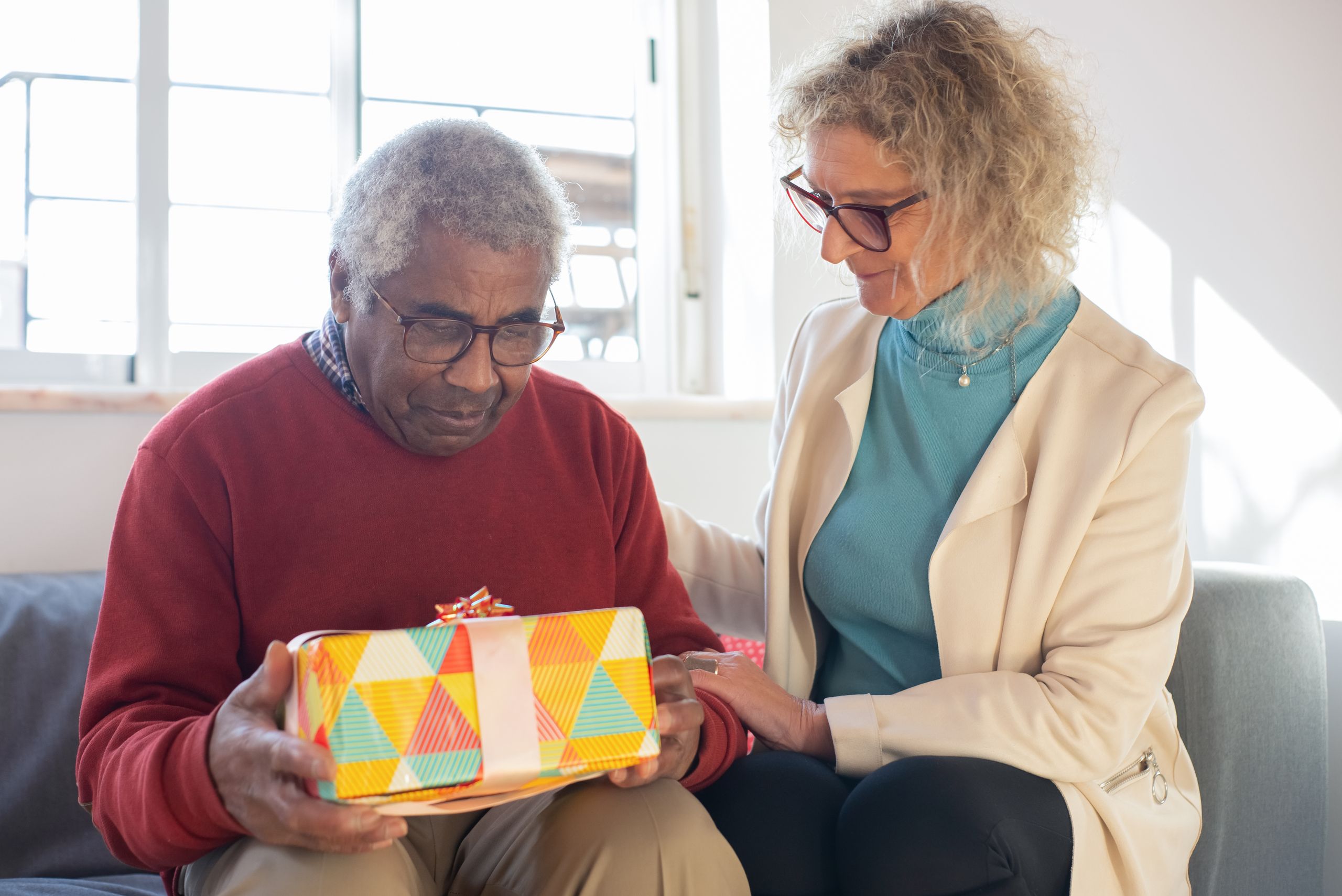 A senior man receives a colorful gift from a caring woman on a couch, indoors with natural light.