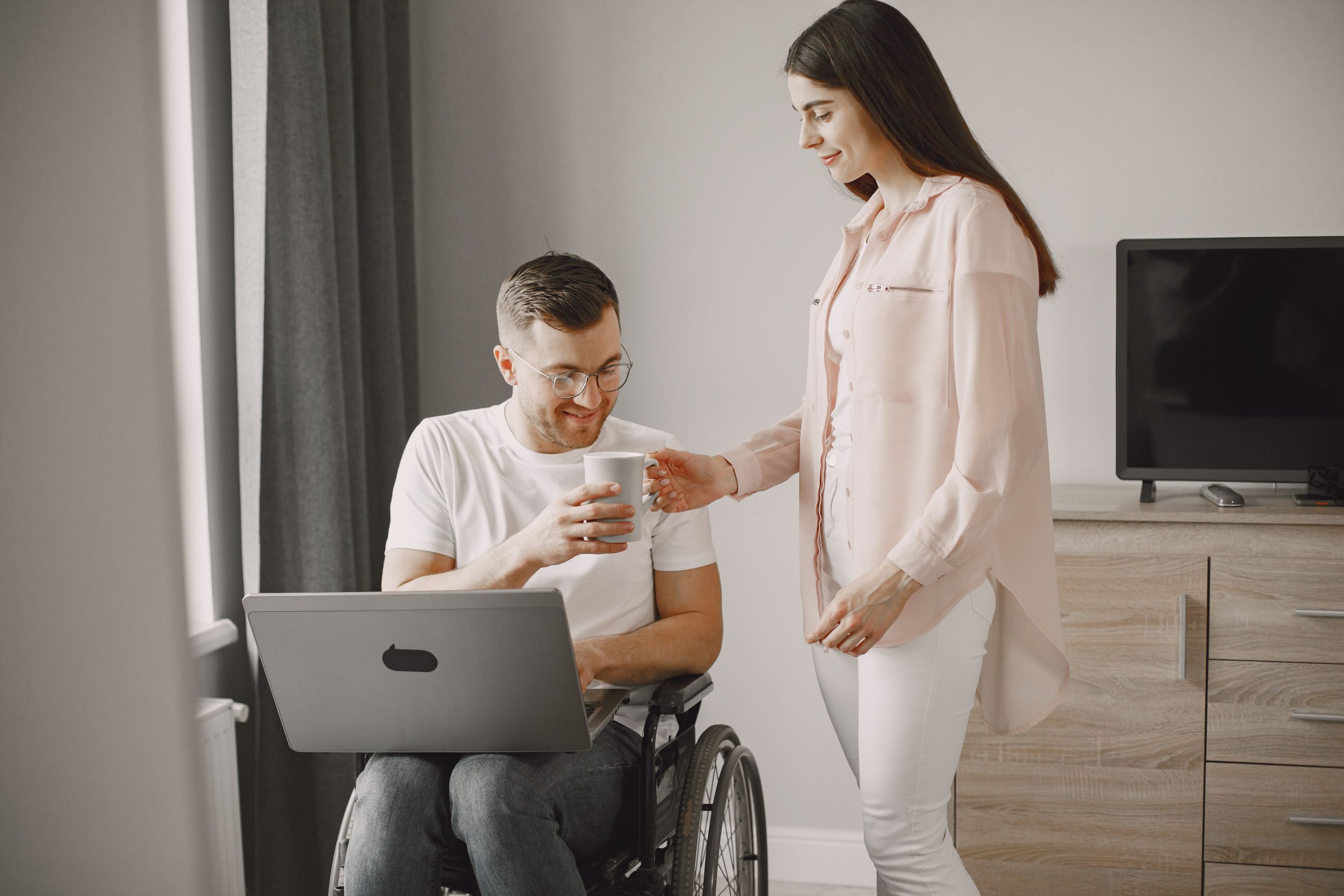 A woman hands a cup to a smiling man in a wheelchair with a laptop, depicting love and care.