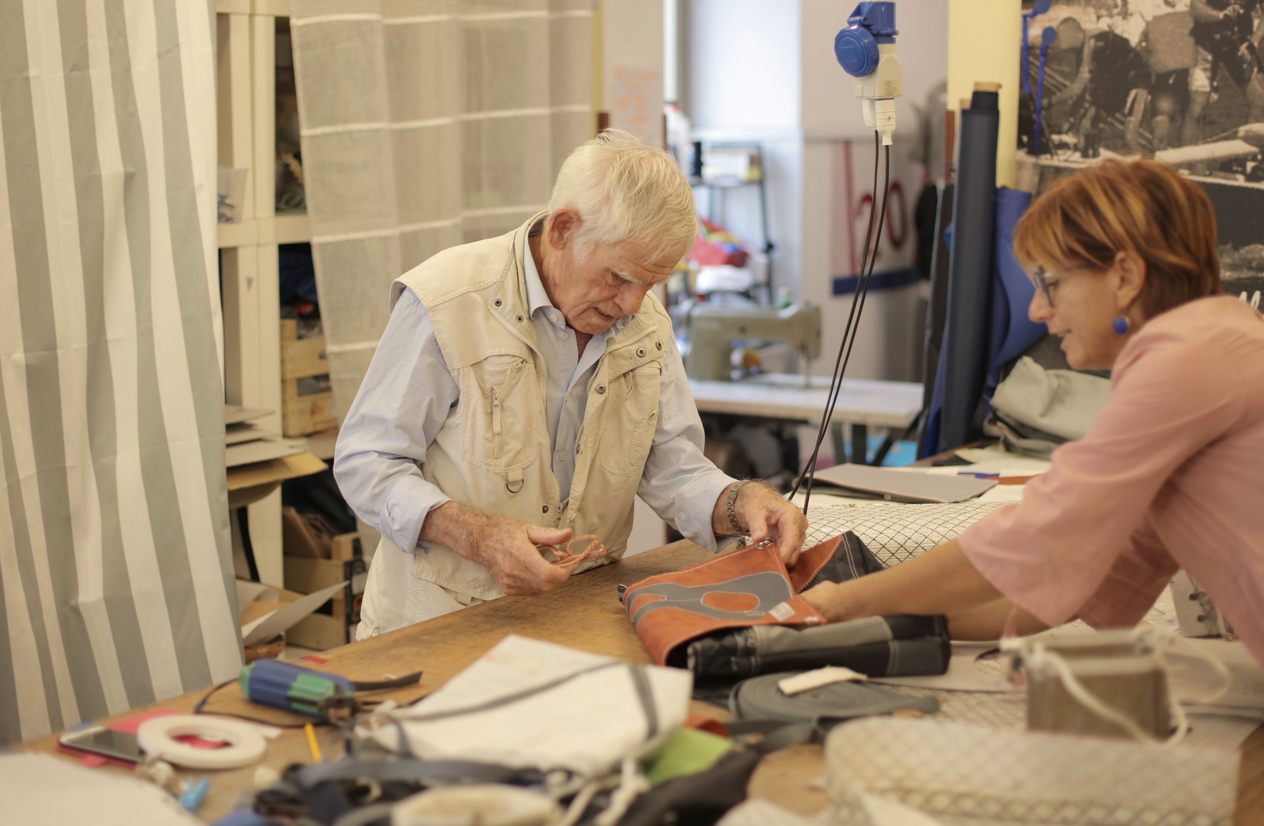 Side view of senior male master in casual wear creating new leather accessory of high quality while standing at table with tools and working together with female partner
